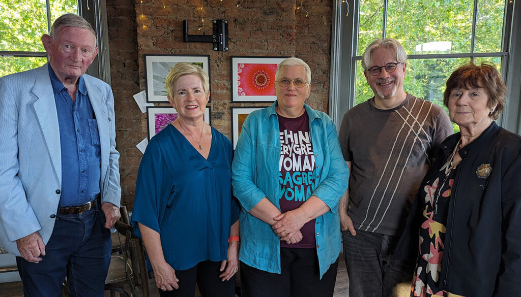 A group of two men and three women standing smiling in front of two windows framing an interior wall on which there are several coloured abstract photographs
