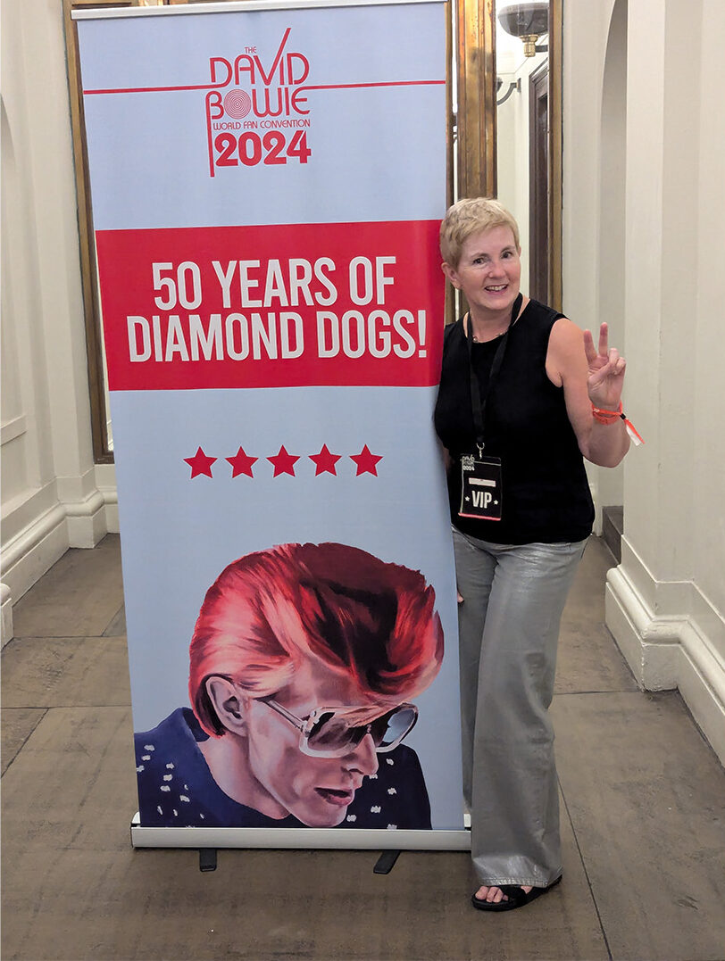 Woman with short hair wearing a black top and sliver jeans next to a banner poster displaying a picture of a man with red hair and sunglasses