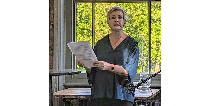A white woman with short blond hair reading from a paper script in front of a sash window with green trees in the background
