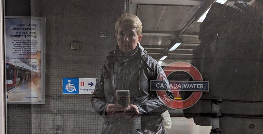 A woman with short blond hair in a long grey coat reflected in a train door in a grey concrete metro station
