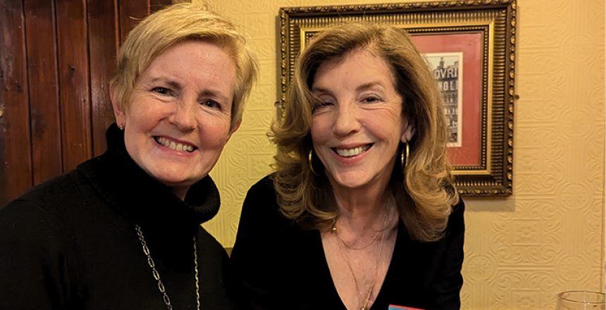 Two women smiling, one with short blond hair, the other with longer dark hair holding a pen to sign a book.